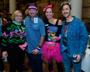 Four smiling people in colorful, themed outfits at a party, with one woman wearing a "New Kids on the Block" shirt.