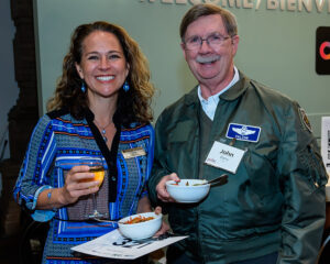 Two people smiling, holding food bowls and drinks, both wearing name tags, one in a blue dress and the other in a green jacket.