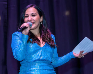 Woman in a blue sparkly dress holding a microphone and a paper, speaking on stage with a dark curtain in the background.