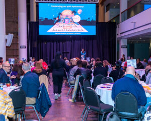 Event with attendees seated at round tables, a speaker on stage, and a screen displaying a welcome message for educators.