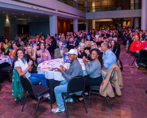 A large, diverse group of people seated at round tables in a spacious hall, smiling and clapping toward the stage.