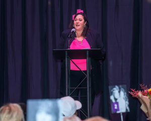 A woman in a pink top and dark blazer speaks at a podium on stage, with a microphone and dark backdrop behind her.