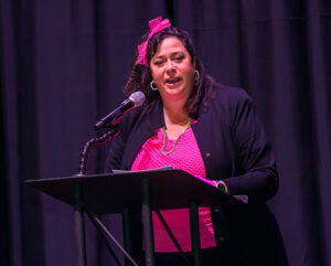 A woman in a pink top speaking at a podium with a microphone on stage.