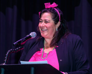 Person wearing pink headband and top, speaking into a microphone on a stand, smiling in front of a dark curtain.
