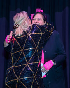 Two people embrace on stage, one wearing a sequined jacket and the other in pink accessories, smiling warmly.