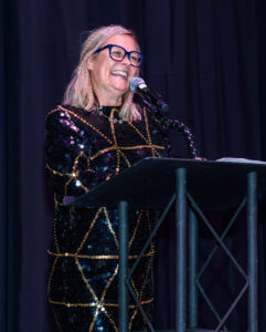 A smiling woman with glasses, wearing a sequin dress, speaks into a microphone at a podium with a dark backdrop.