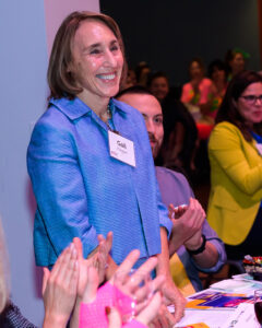 A smiling person with a name tag stands while others around them clap at an indoor event.