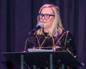 A person with glasses and blonde hair speaks into a microphone while standing at a podium on a stage.