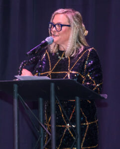 Person with glasses and a sequined dress speaking at a podium on a stage with a dark backdrop.