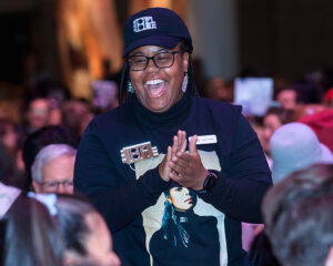 Person clapping and smiling at an event, wearing a black cap and a black shirt with a photo of a woman on it.