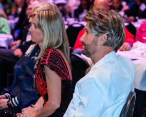 People sitting and attentively listening at a conference, with focus on a man in a white shirt and a woman in a red and black outfit.