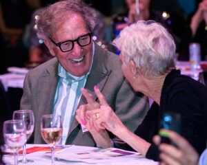 An elderly man and woman are laughing and talking at a table with wine glasses.
