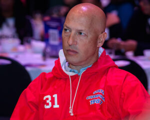 A bald man wearing a red jacket with the text "State Champion" and the number 31 sits at a table, looking serious.