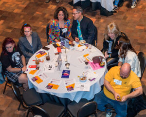 A group of people sit around a round table at an event, some engaged in conversation, with papers and drinks on the table.