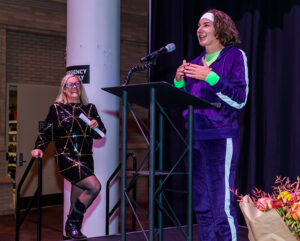 Two women on stage; one speaks at a podium in a velvet tracksuit, the other smiles and stands in a sequined dress.