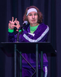 Person in a purple tracksuit and headband speaks at a podium, making a gesture with one hand.