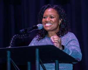 A person smiling and speaking into a microphone at a podium, wearing a sparkly long-sleeved top.