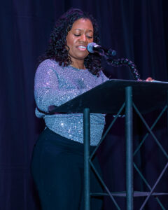 A person speaks at a podium with a microphone, wearing a sparkly top and dark pants, with a dark curtain in the background.