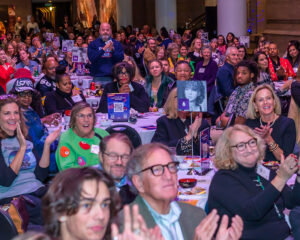 A diverse group of people clapping and smiling while seated at a crowded indoor event.