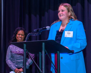 Two women at an event, one in a blue suit speaking at a podium, and another in a sparkly outfit standing in the background.