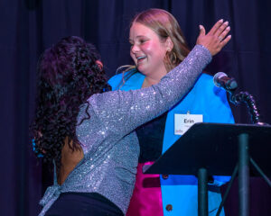 Two women smiling and embracing on stage, one wearing a sparkly top and the other in a blue blazer with a name tag reading "Erin".