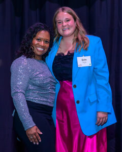 Two women smiling, standing close together; one in a glittery top and black skirt, the other in a blue blazer and pink skirt.