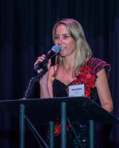 A woman in a formal black and red dress speaks into a microphone on stage, with a name tag that reads "Kendee Rank".