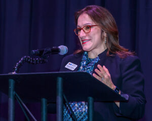 A woman with glasses speaks into a microphone at a podium, smiling and gesturing with her hands.