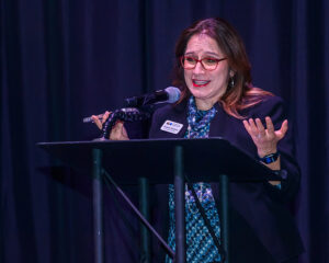 A woman with glasses speaks into a microphone at a podium, gesturing with her hands against a dark curtain backdrop.