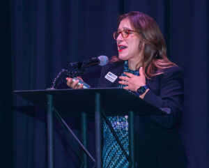 A person speaks passionately at a podium with a microphone, wearing glasses and a name tag, hand on chest.