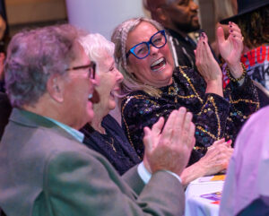 Three people sit at a table, laughing and clapping, while enjoying an event.