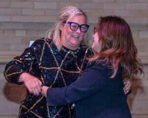 Two women with glasses embrace and smile in front of a brick wall.