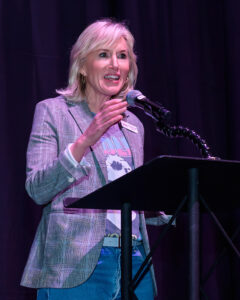 A woman with blonde hair speaks into a microphone while standing at a podium, wearing a blazer and jeans.