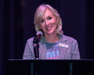 A woman in a blazer speaking at a podium with a microphone, smiling, and wearing a name tag.