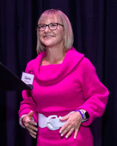 A woman in a bright pink sweater and glasses smiles, standing in front of a dark background. She's wearing a name tag.