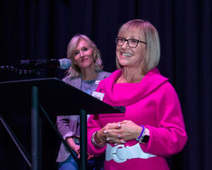 Two women are at a podium; one speaks and smiles in a pink outfit, while the other stands behind her and listens.