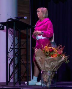 Person in a bright pink outfit speaks at a podium with a bouquet of flowers nearby on stage.