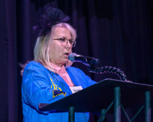 A woman standing at a podium, speaking into a microphone, wearing a blue outfit and glasses with a name tag reading "Susan.