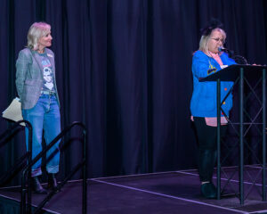 Two women on stage with black curtains; one speaks at a podium while the other stands to the side holding papers.