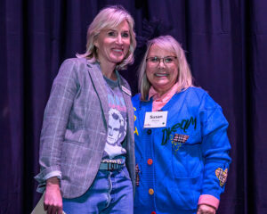 Two women smiling and posing together, one wearing a plaid blazer and graphic tee, and the other in a blue cardigan.