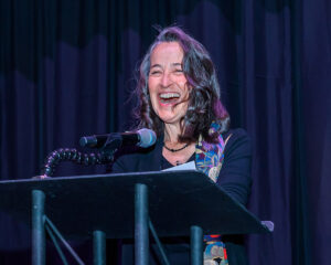 A woman speaking and smiling at a podium with a microphone in front of a dark curtain.