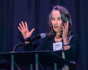 A woman speaking at a podium with a microphone, raising both hands. Name tag reads "Susan.