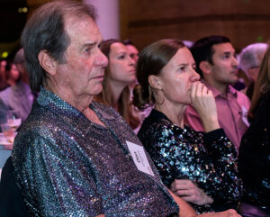 A man and woman in sequined outfits sit attentively among a group of people at an indoor event.