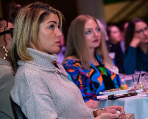 A group of attentive women sit at a table during a conference, with one woman in focus wearing a turtleneck sweater.