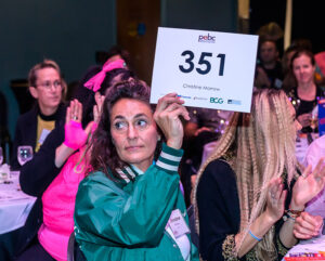 A woman in a green jacket holds up a bidding sign numbered 351 at an event, with people clapping in the background.