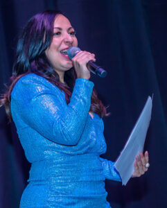 Woman in a blue dress holding a microphone and papers, speaking on stage.