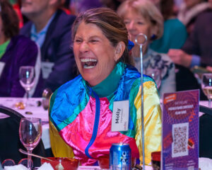 A woman wearing a colorful jacket laughing at a formal event, seated at a table with a name tag that reads "Molly.