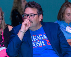 Man in glasses wearing a blue blazer over a "Reagan Bush '84" t-shirt, sitting with his hand over his mouth.