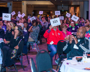 People at an auction event hold up numbered paddles while seated at round tables in a large room.