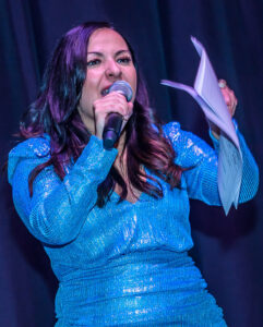 A person in a sparkling blue dress passionately speaks into a microphone while holding papers on stage.